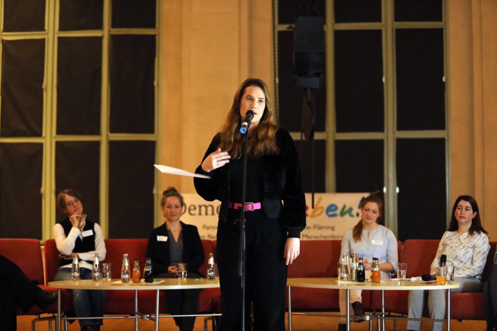 Eine Frau steht in der Mitte der Bühne und trägt ein Spoken Words Beitrag vor. Im Hintergrund sitzen Podiumsteilnehmerinnen